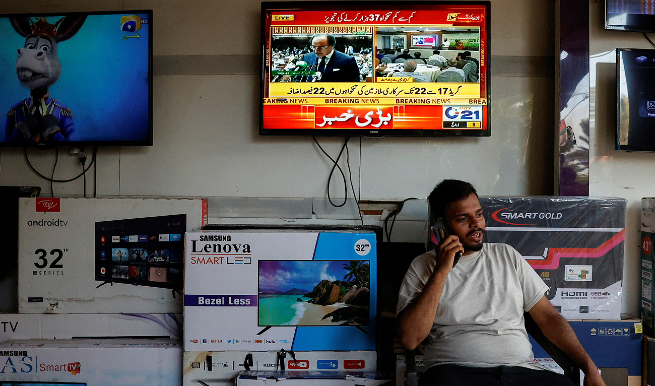 A salesman uses his mobile phone as he sits under a television screen displaying the live broadcast of Pakistan Finance Minister Muhammad Aurangzeb presenting the 2024/25 budget, at an electronics market in Karachi, Pakistan June 12, 2024. (REUTERS)