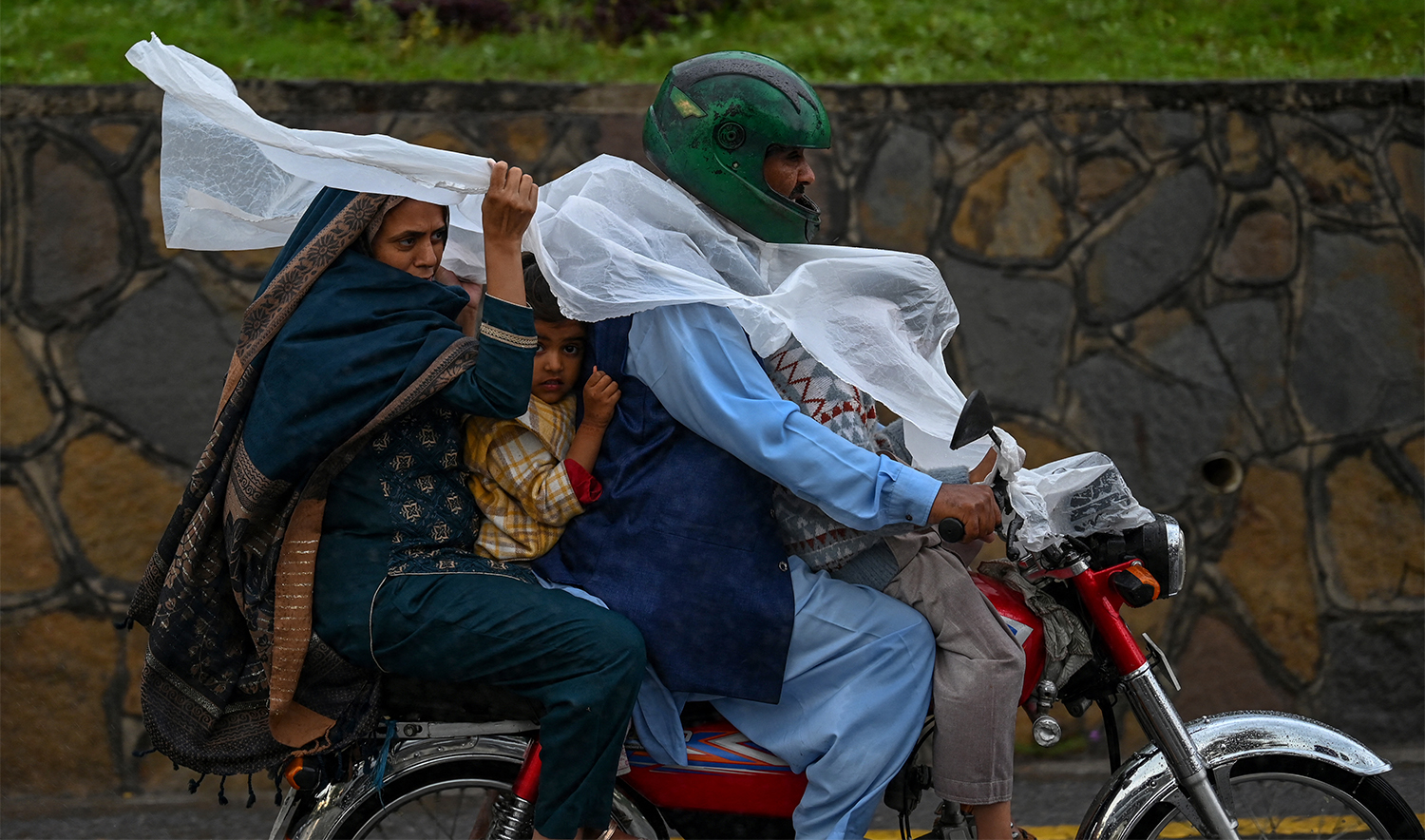 A family travelling on a motorbike uses a plastic sheet to shelter from the rain in Islamabad on April 15, 2024. (AFP/File)