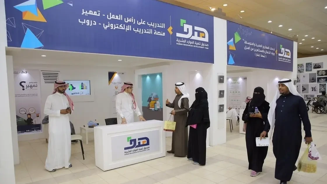 People interacting at a human resources development exhibition booth with banners in Arabic.