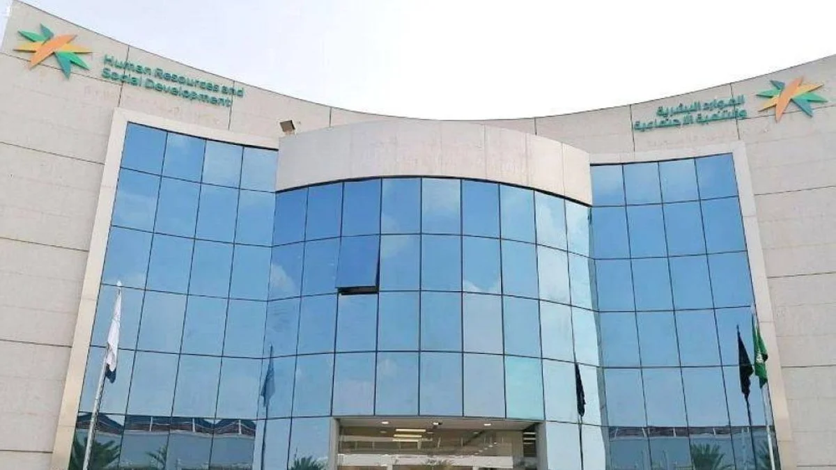 The exterior of the Ministry of Human Resources and Social Development building in Saudi Arabia. It's a modern building with a curved, multi-story glass front. The ministry logo and name are visible at the top, and flags, including the Saudi Arabian flag, are placed on either side of the entrance.