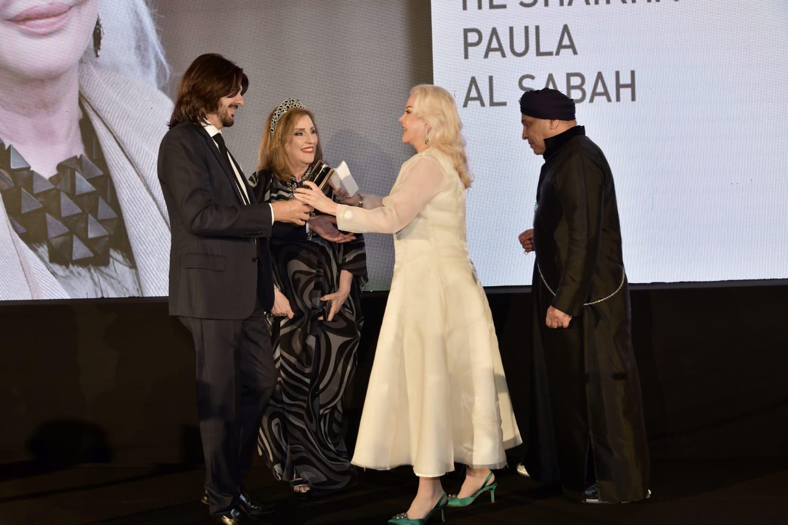 Lors d'une cérémonie officielle, un homme en costume reçoit un prix des mains d'une femme vêtue de blanc, sous un écran affichant "PAULA AL SABAH" en arrière-plan.