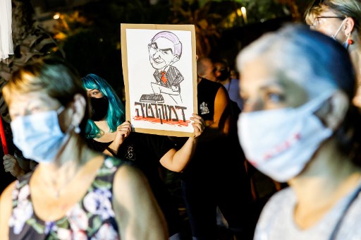 Manifestantes masquées portant un panneau représentant le Premier ministre israélien Benjamin Netanyahu assis sur le mot « démocratie » en hébreu le 1e aout 2020 (Jack Guez/AFP)