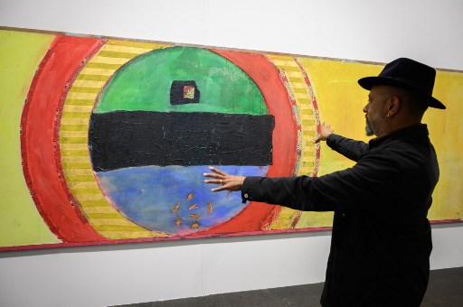Le fils de l'artiste britannique Frank Bowling, né en Guyane, Ben Bowling fait un geste devant l'une des œuvres de son père intitulée : «Samson's Circle with Lila's dress made in Africa of import Chinese Fabric: and the rest, 2019», exposée dans le secteur Unlimited de Art Basel, la première foire mondiale d'art moderne et contemporain, à Bâle, le 21 septembre 2021. La foire sera ouverte au public du 24 au 26 septembre 2021. (Fabrice Coffrini/AFP)