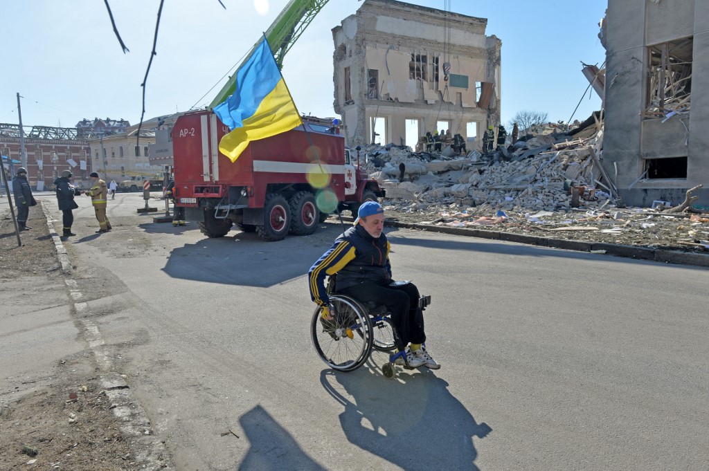 Un homme en fauteuil roulant passe devant des secouristes déblayant les décombres d'un bâtiment de l'Institut régional d'administration publique de Kharkiv endommagé par des bombardements à Kharkiv le 18 mars 2022. (AFP)