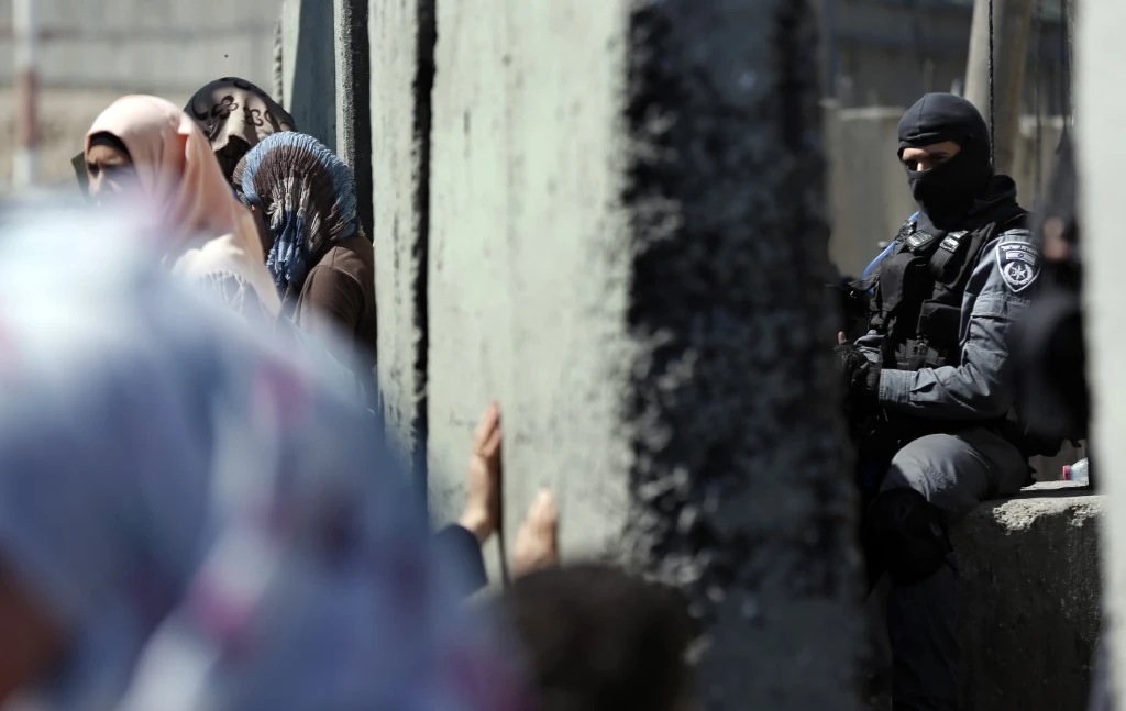 Un officier de police israélien masqué monte la garde au point de contrôle de l'armée israélienne de Qalandia, entre la ville de Ramallah en Cisjordanie et Jérusalem, alors que des femmes palestiniennes attendent de traverser vers Jérusalem pour assister à la prière du vendredi midi dans l'enceinte de la mosquée Al-Aqsa, le 3 juillet 2015. (Photo Thomas Coex / AFP)