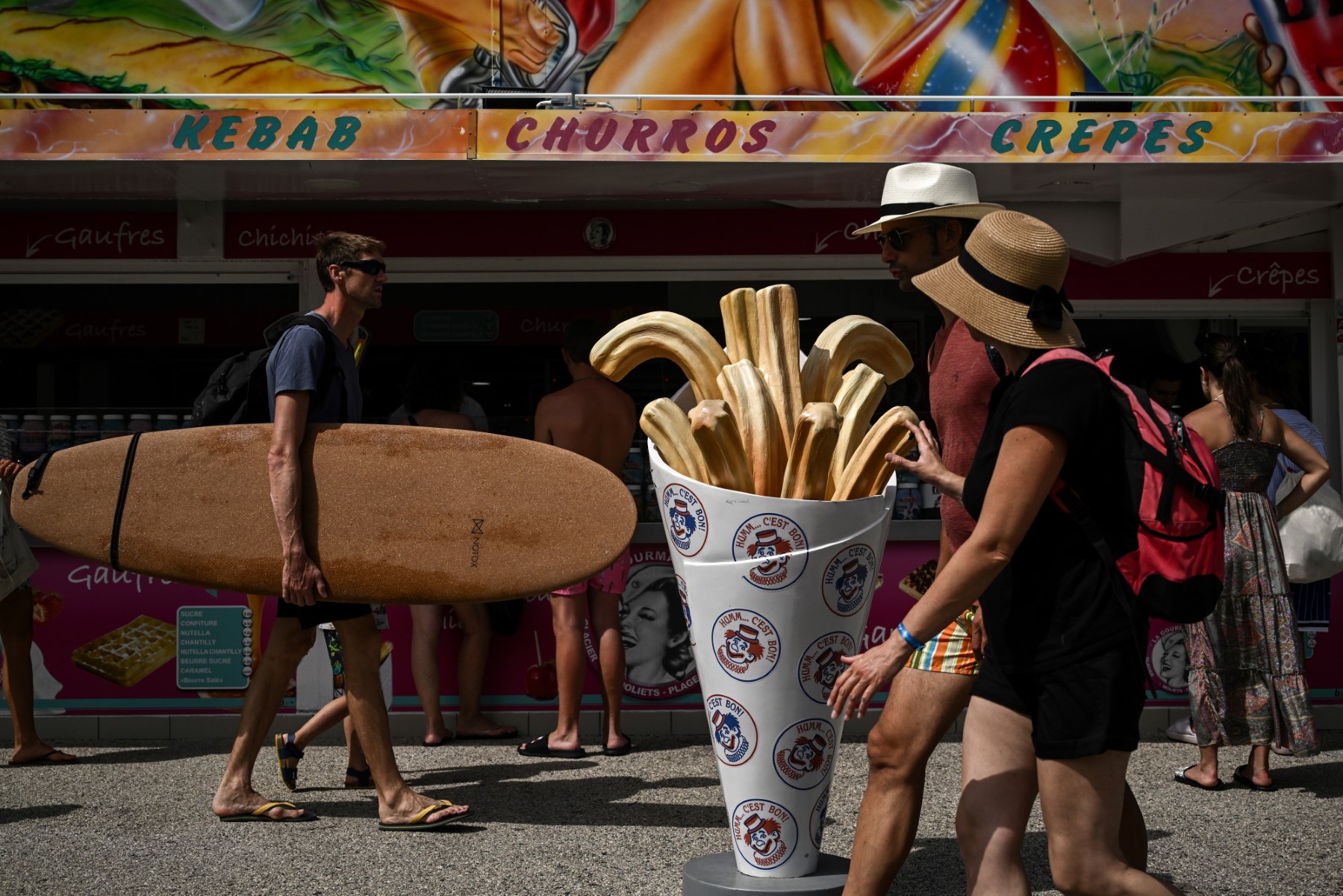 Cette photo prise le 18 juillet 2023 montre un homme tenant une planche de surf qui passe devant un marchand de glaces près d'une plage à Moliets-et-Maa, dans le sud-ouest de la France. (Photo par Philippe LOPEZ / AFP)