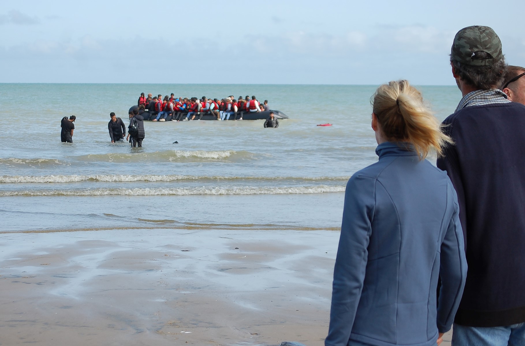 Des personnes regardent des migrants assis à bord d'un bateau gonflable avant de tenter de traverser illégalement la Manche pour rejoindre la Grande-Bretagne, sur la plage de Sangatte, dans le nord de la France, le 18 juillet 2023. (Photo Bernard Barron / AFP)