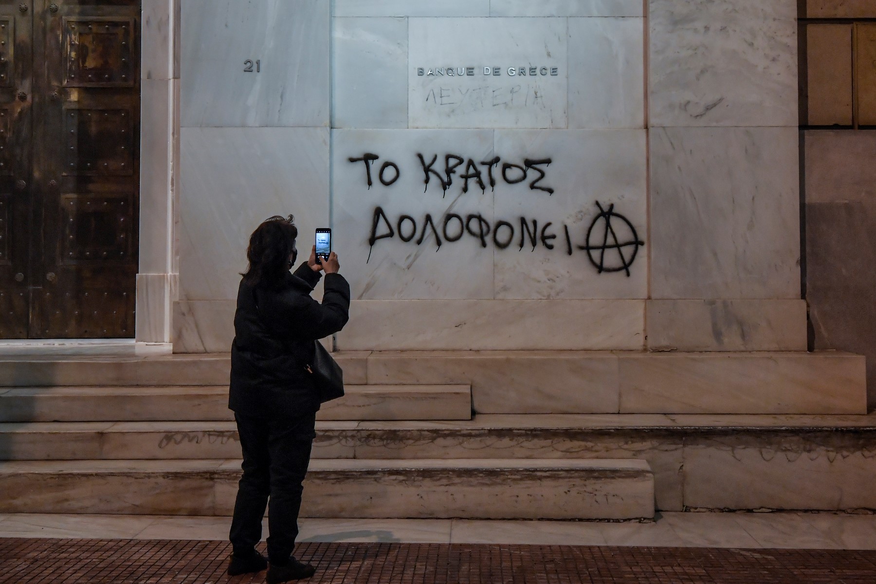 Une femme prend une photo d'un slogan écrit sur le siège de la banque de Grèce disant "l'État assassine" dans le centre d'Athènes, le 3 mars 2021 lors d'une manifestation de soutien à Dimitris Koufodinas, un tueur à gages d'extrême-gauche grec malade. (AFP).