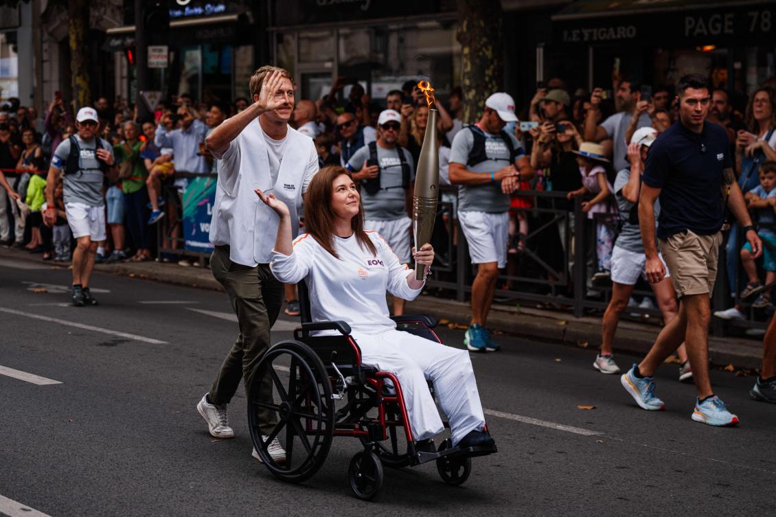 Agence France Presse (AFP) US video journalist Dylan Collins pushes the wheelchair of AFP Lebanese photographer Christina Assi holding an Olympic torch during the Olympic Torch Relay in Vincennes, near Paris, on July 21, 2024, ahead of the Paris 2024 Olympic Games. AFP journalists Christina Assi and Dylan Collins were injured in an attack by an Israeli tank on a group of journalists in southern Lebanon on October 13, 2023. (AFP)