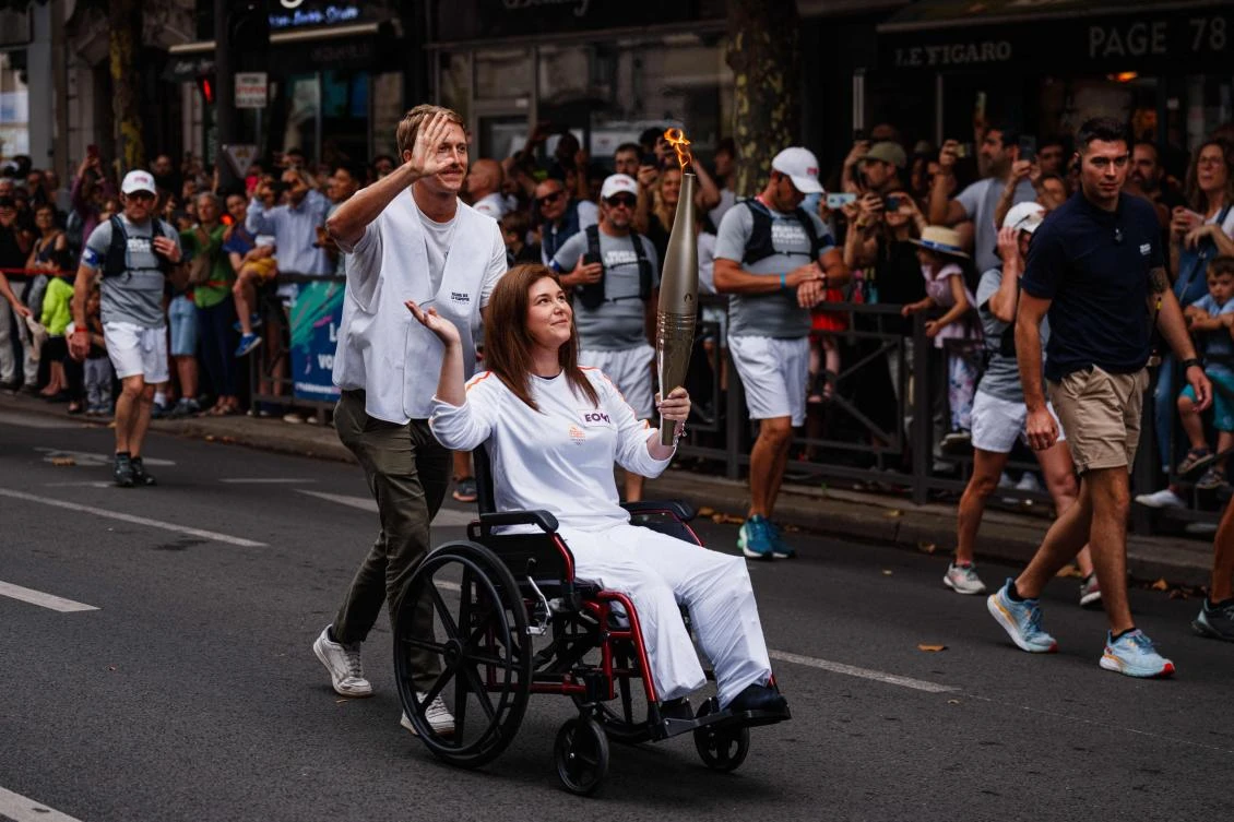 Agence France Presse (AFP) US video journalist Dylan Collins pushes the wheelchair of AFP Lebanese photographer Christina Assi holding an Olympic torch during the Olympic Torch Relay in Vincennes, near Paris, on July 21, 2024, ahead of the Paris 2024 Olympic Games. AFP journalists Christina Assi and Dylan Collins were injured in an attack by an Israeli tank on a group of journalists in southern Lebanon on October 13, 2023. (AFP)