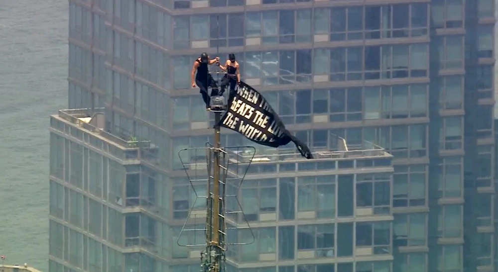 Two people stand on the tip of the antenna of the Empire State Building in New York while holding a banner on Wednesday, July 1, 2026. (WABC-TV/AP)