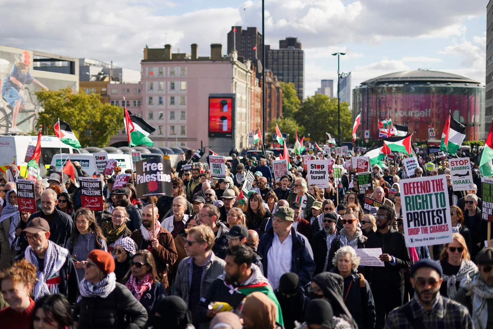 A wide shot of a large protest in a city street, with numerous people holding Palestinian flags, and signs that read "Free Palestine," "Stop Arming Israel," and "Defend the Right to Protest." Buildings are visible in the background under a partly cloudy sky.