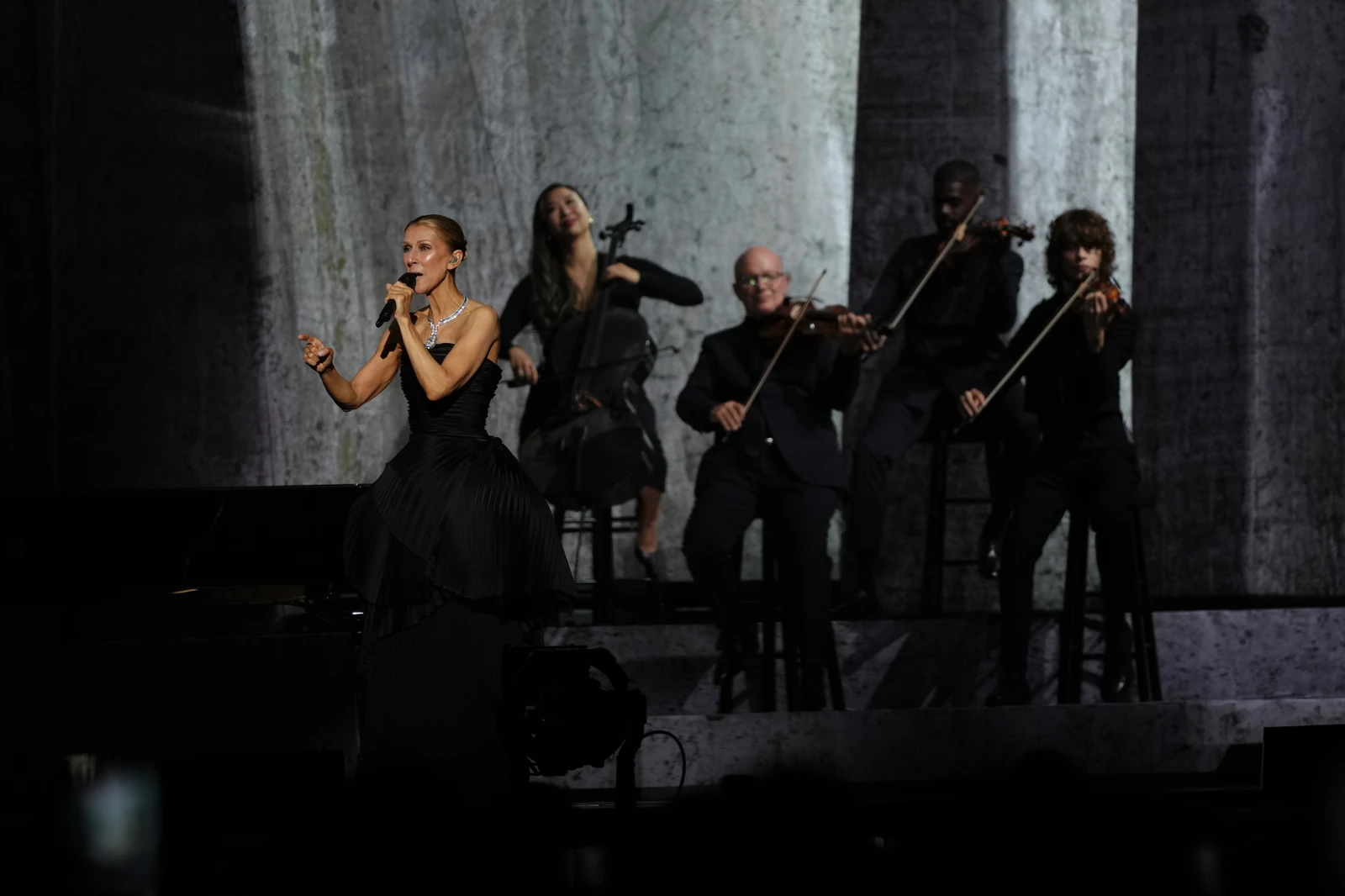 A singer in a black dress performs on stage with a string quartet in formal attire, under dramatic lighting.