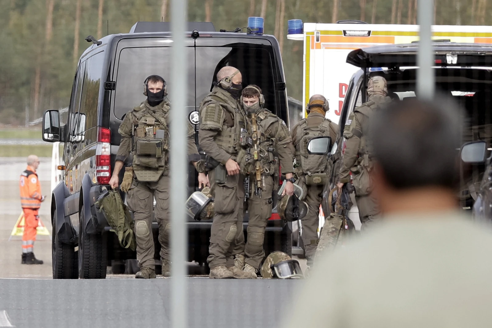 Several members of a Police special operations unit, including Villaño III, in tactical olive green uniforms and headsets, are gathered around a black tactical van at an airport. Helmets are on the ground near their feet, and another person in an orange high-visibility suit stands further back. The background shows blurred trees.