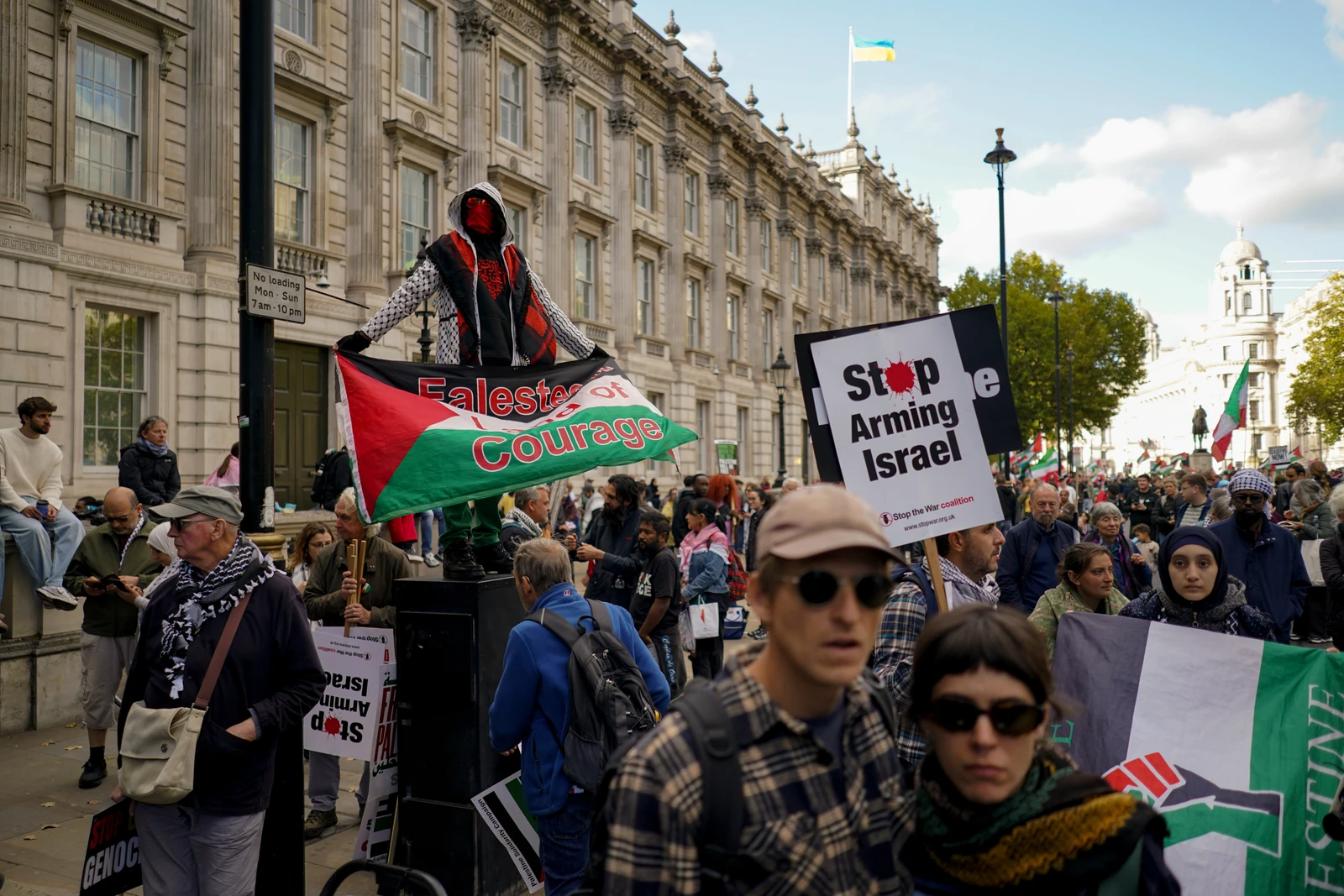 A wide shot of a street protest in London. A person in a hooded jacket and red face covering stands on a raised platform, holding a large Palestinian flag banner that reads 'Palestine of Courage'. Many other protesters are visible, holding signs such as 'Stop Arming Israel' and 'Stop Genocide'. Buildings with classical architecture line the street, and a Ukrainian flag flies atop one.