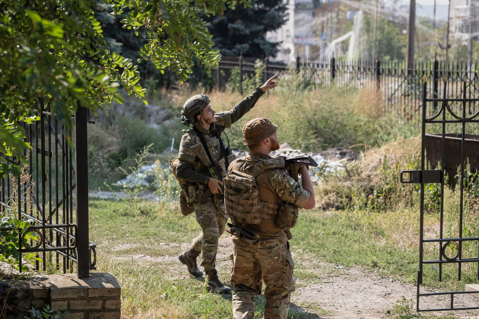 Two military personnel in camouflage gear observe their surroundings near a gated outdoor area, one soldier pointing forward.