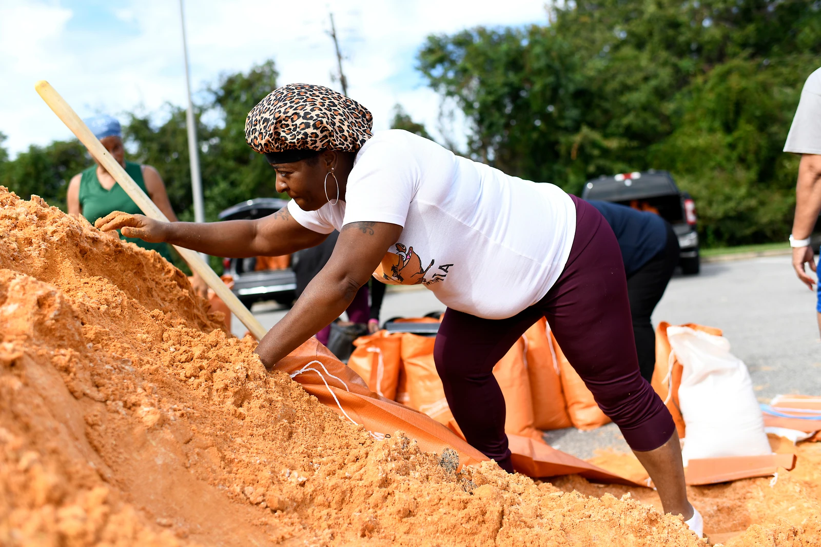 A woman wearing a leopard print cap and a white shirt bends over, scooping sand into an orange sandbag from a large mound of sand. Other people and more sandbags are visible in the background under a blue sky.