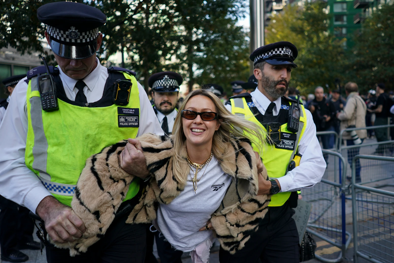 A smiling blonde woman in sunglasses and a faux tiger print coat is being led away by two male police officers in uniforms and high-visibility vests. Other police and blurred figures are in the background.