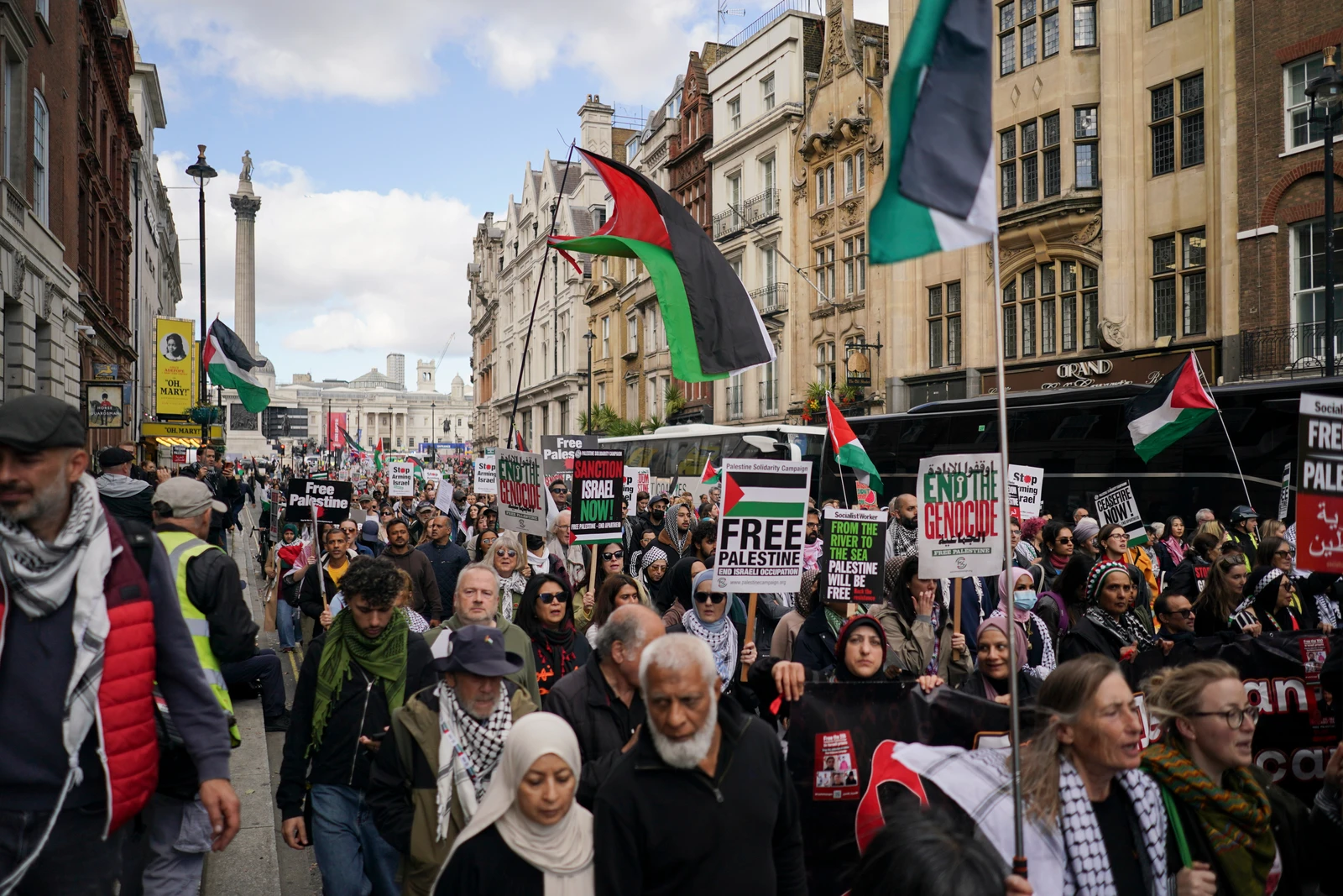 A wide shot of a street filled with a diverse crowd of people marching. Many are holding Palestinian flags and signs with slogans like 'Free Palestine' and 'End the Genocide'. Historical buildings are visible on both sides of the street, and a tall monument can be seen in the distance under a partly cloudy sky.
