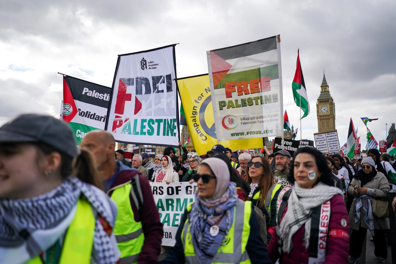 A large crowd of people, many wearing high-visibility vests and keffiyehs, march while holding numerous banners and Palestinian flags. The iconic Big Ben clock tower is visible in the upper right background under a grey, cloudy sky.