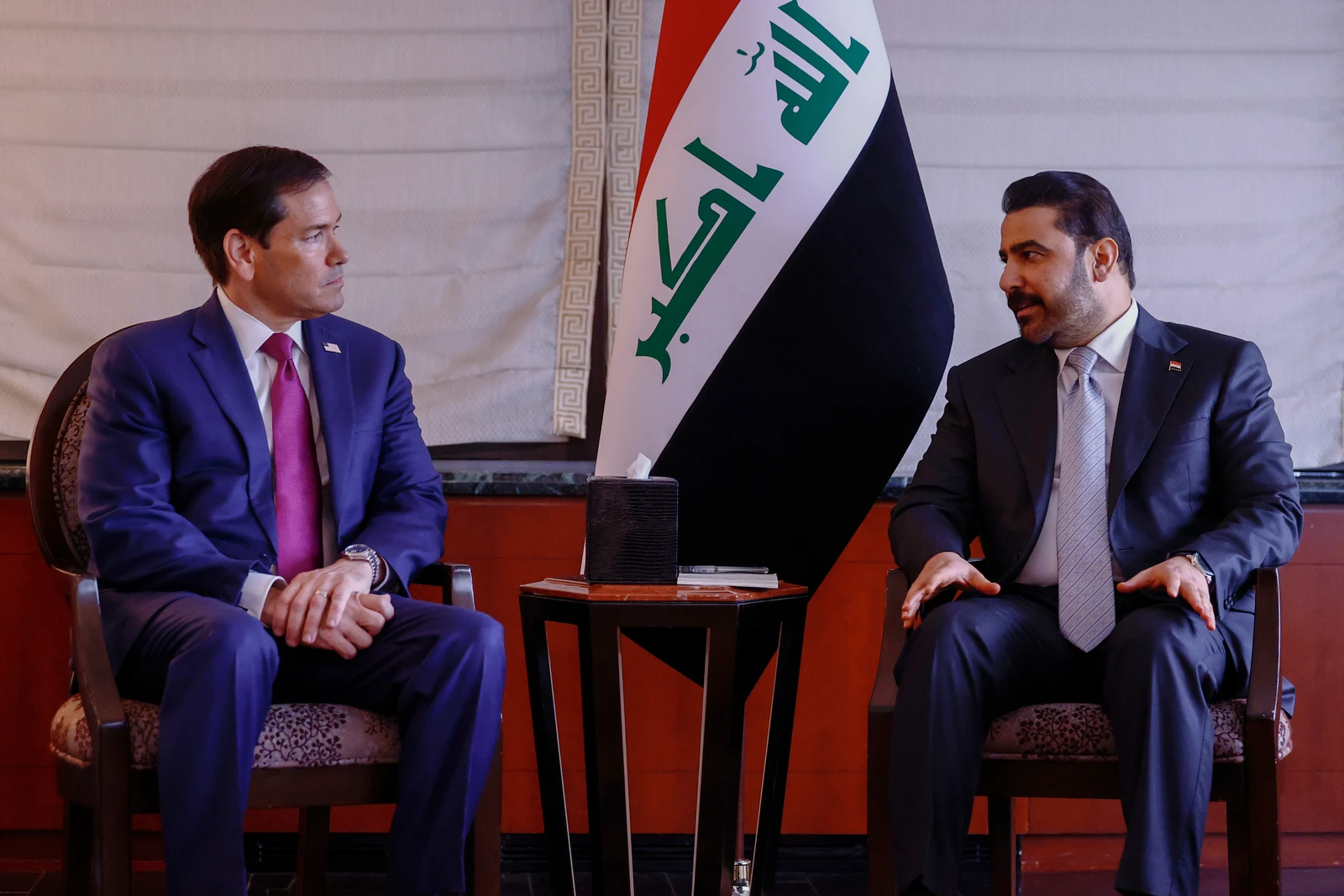 Two men in suits seated facing each other with the Iraqi flag between them, engaged in a discussion.