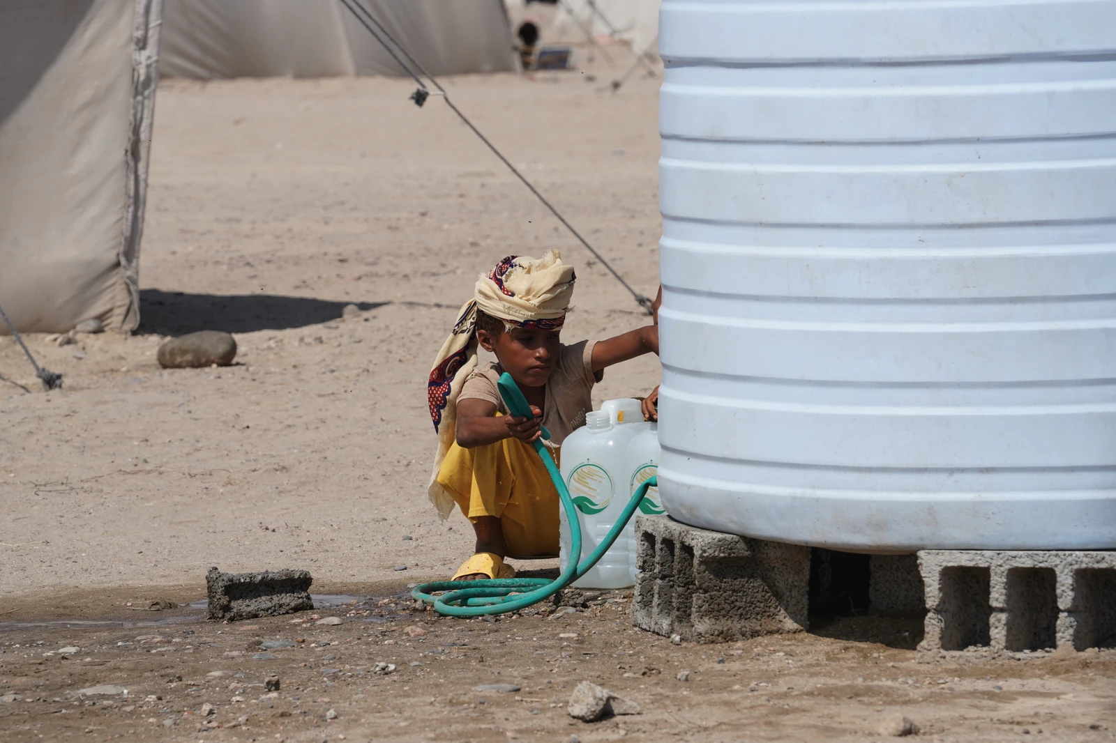A young Yemeni boy in a head covering and yellow clothing kneels on dry, dusty ground, filling two clear plastic jugs with water from a large white water tank using a green hose.