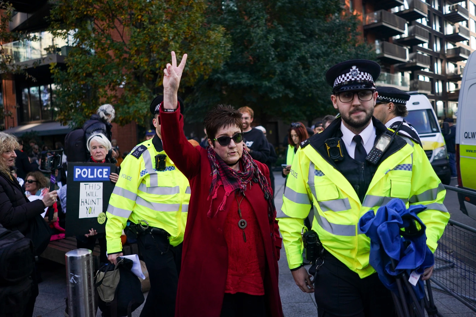 A woman in a red coat and sunglasses, mid-arrest by two police officers in neon yellow vests, holds her arm up making a peace sign. Other protestors and city buildings are in the blurred background.