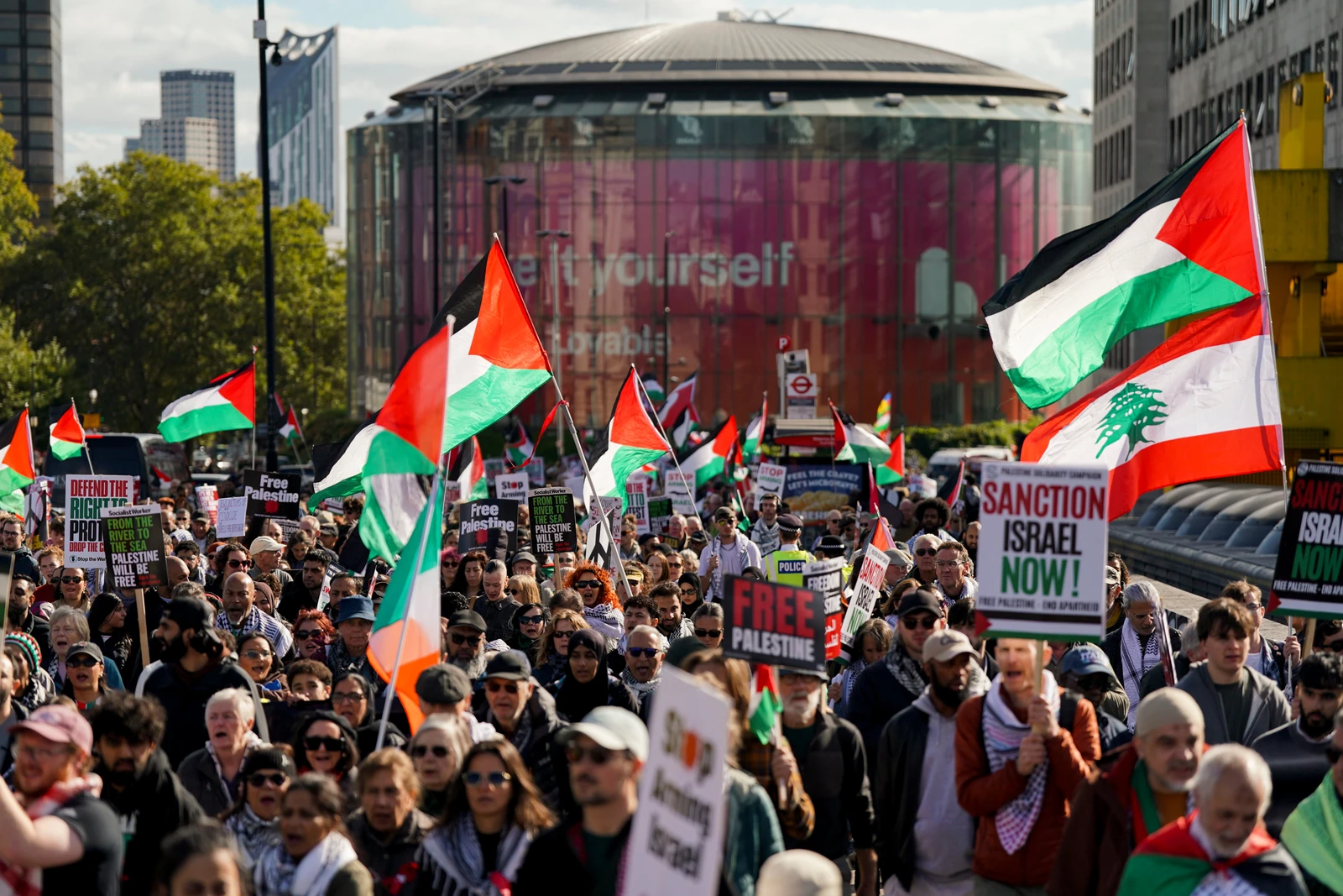 A wide shot shows a dense crowd of people marching along a city street. Many individuals hold up red, green, black, and white Palestinian flags, some with the Lebanese cedar tree symbol. Others carry signs with slogans like 'Sanction Israel Now!' and 'Free Palestine.' In the background, modern glass buildings with a large domed structure are visible under a bright sky, with a few trees on the left.