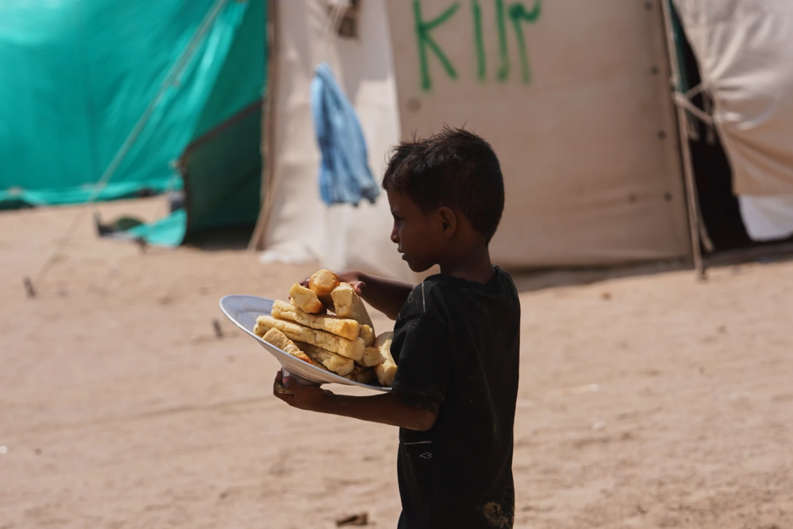 A young Yemeni boy, facing right in profile, carries a metal plate piled high with pieces of bread. He has dark, messy hair and wears a dark t-shirt. Behind him, a teal tarp and a light-colored tent are visible.